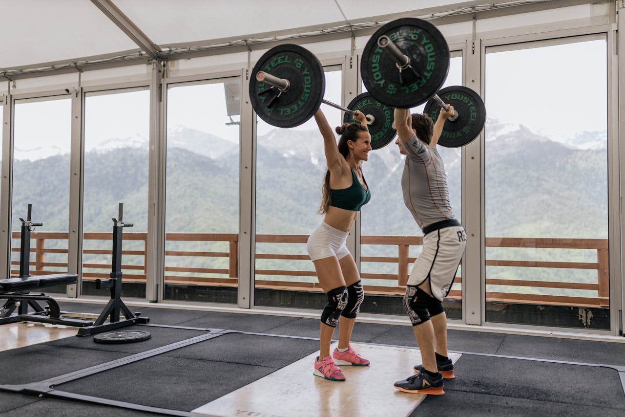 A couple lifting barbells together in a gym with scenic mountain views.