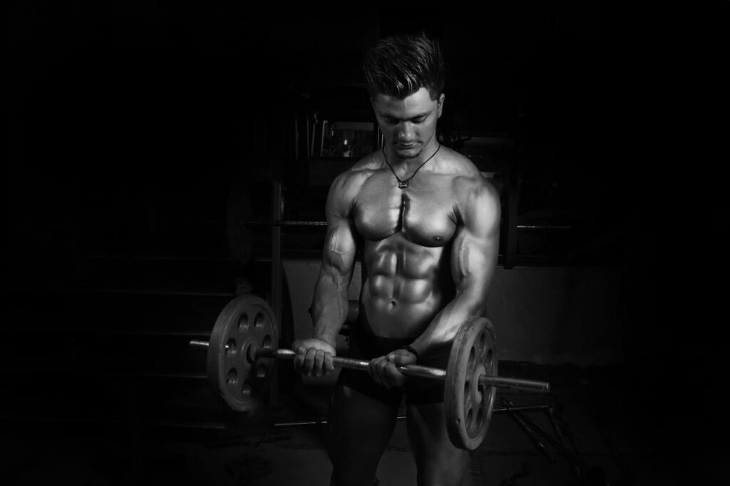 Shirtless male bodybuilder lifting barbell in a dark gym, emphasizing muscles.